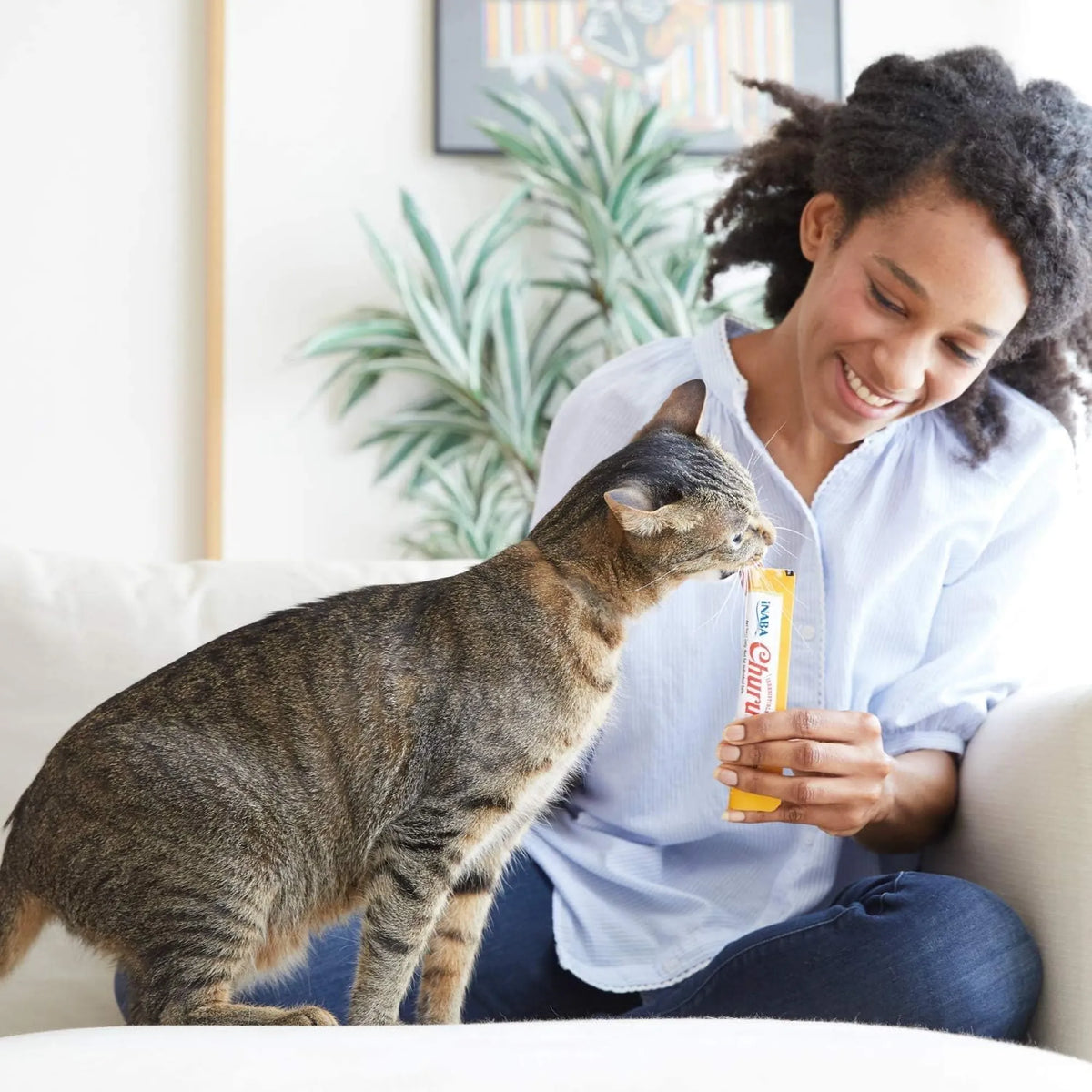 Mujer sonriente dando un tubo de puré a un gato atigrado en el salón de casa