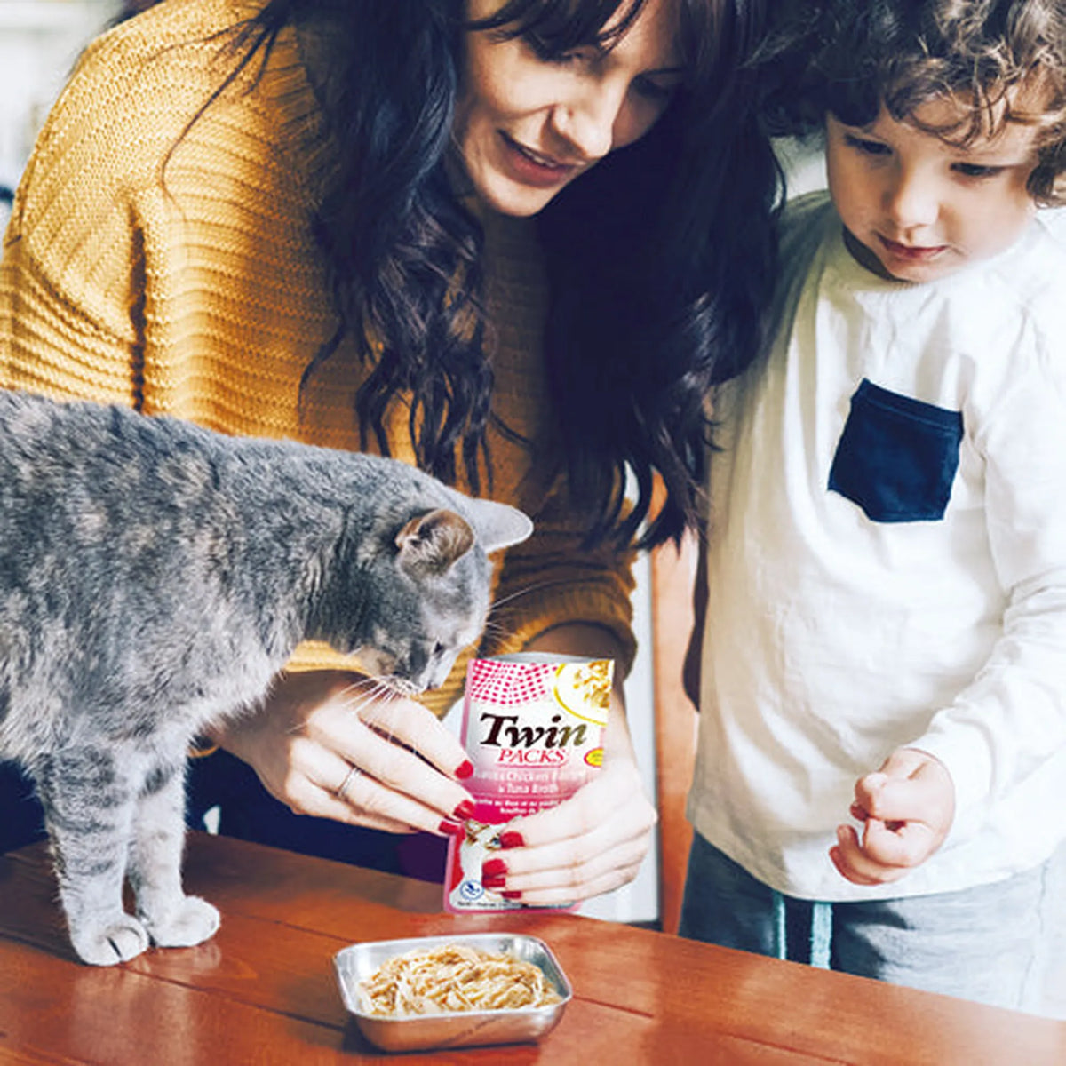 Una mujer y un niño dan de comer a un gato gris en la cocina con una bolsita recién abierta